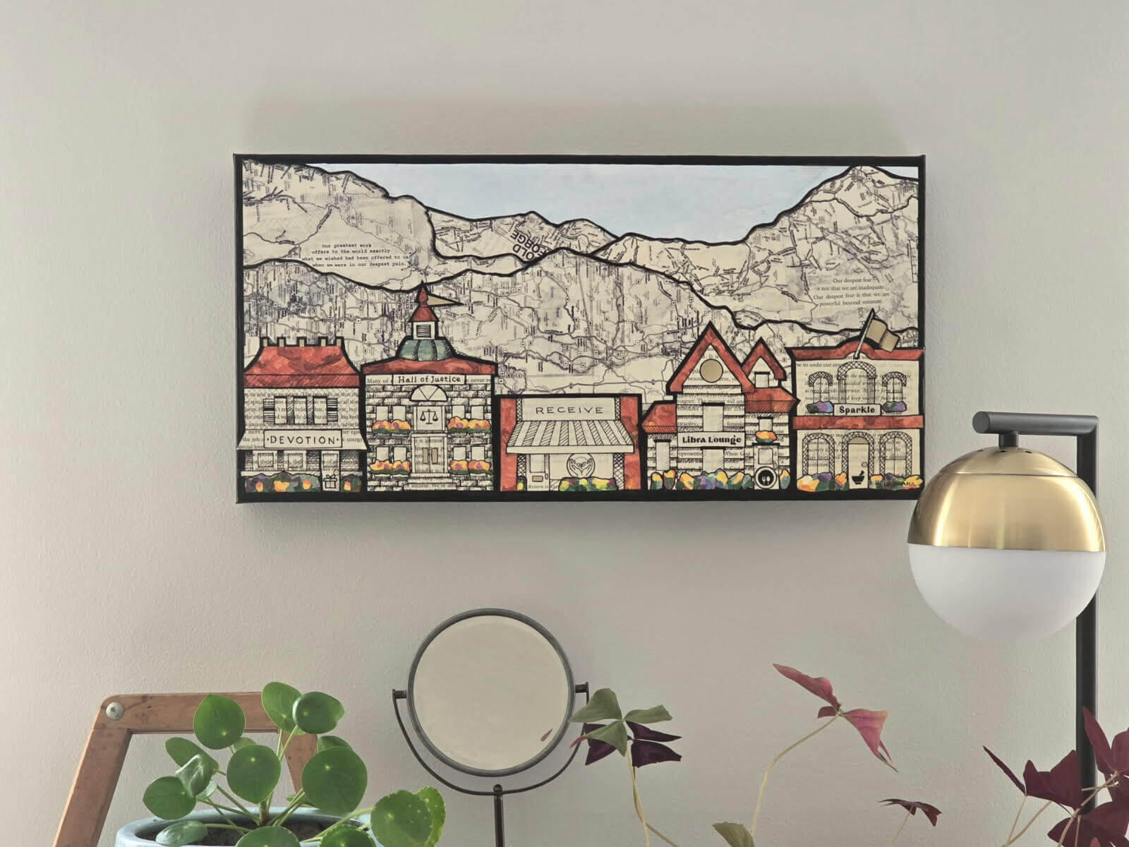 Lisa Hunter's collage consisting of a row of hand drawn shops and houses with orange yellow purple and green accents in front of mountain ranges and blue sky containing words and maps printed on book pages outlined in black. Feather Road Design Studio, LLC