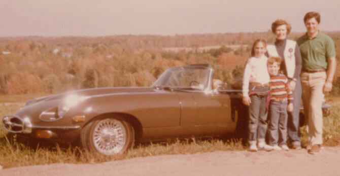 Lisa Hunter's vintage, faded color photo, 1970s, family of four next to classic brown convertible Jaguar on a dirt road. Fall background. Feather Road Design Studio, LLC