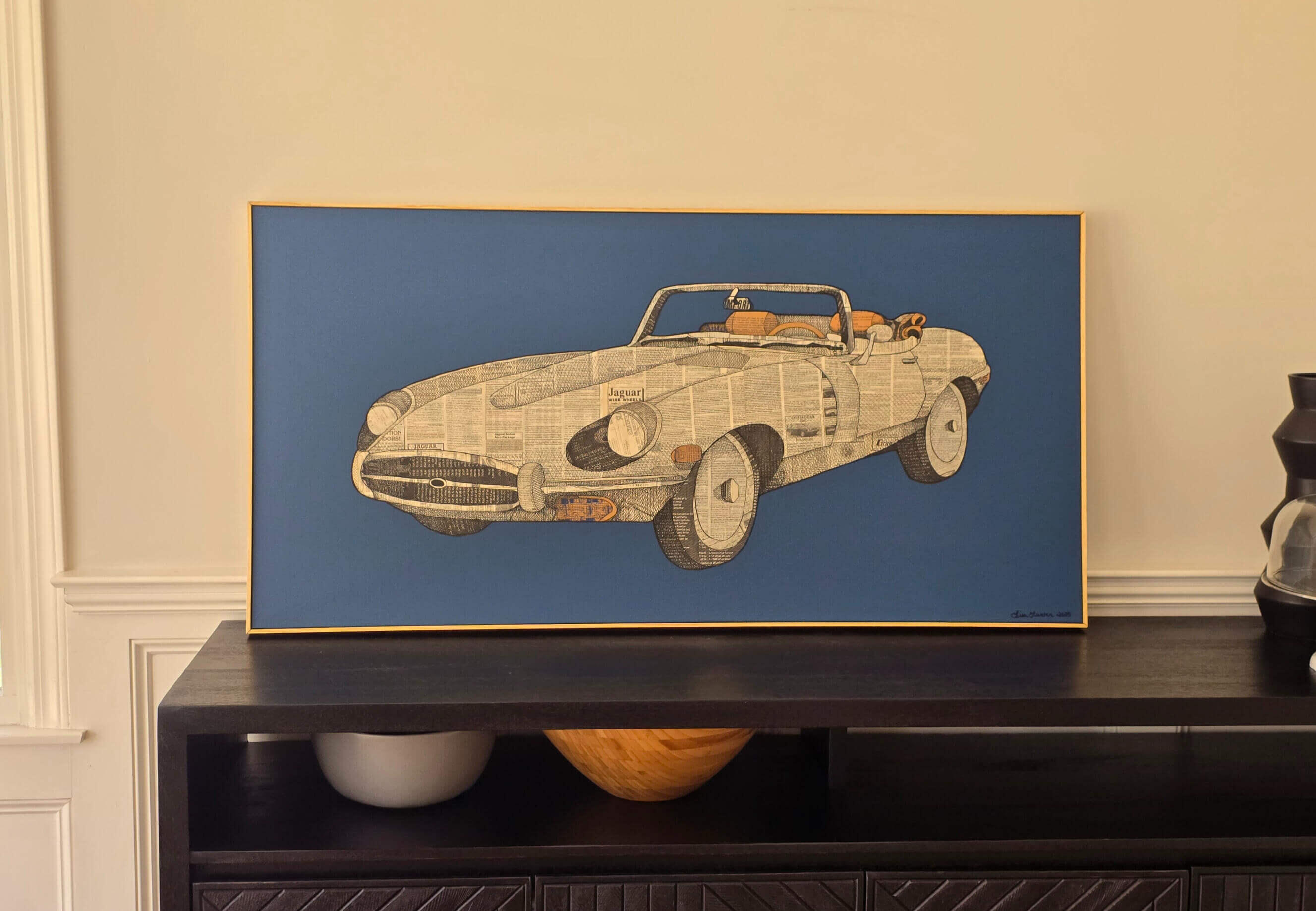 Lisa Hunter' wood framed horizontal rectangular artwork sitting on black table. Side view of vintage convertible Jaguar sports car collage, vintage black and white swap sheet ads, burnt umber accents on blue background, Feather Road Design Studio, LLC